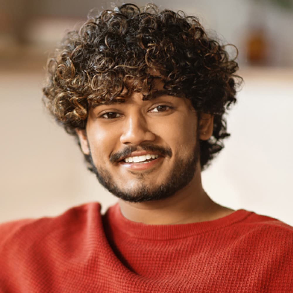 A smiling man with curly hair and confidently with her arms crossed, wearing a bright orange shirt. The background features soft, neutral tones, suggesting a warm and welcoming environment.