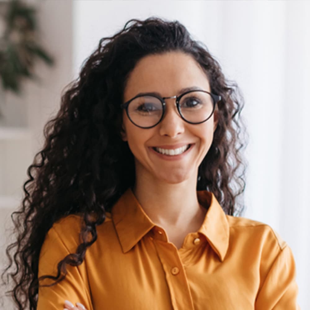 A smiling woman with curly hair and glasses stands confidently with her arms crossed, wearing a bright orange shirt. The background features soft, neutral tones, suggesting a warm and welcoming environment.